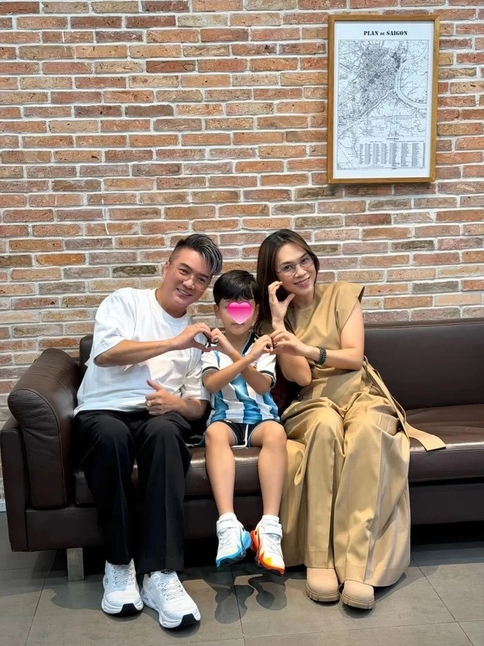 Mỹ Tâm mặc váy vàng ngồi trên sofa đùa với bé Polo bên cạnh Đàm Vĩnh Hưng trước bức tường gạch